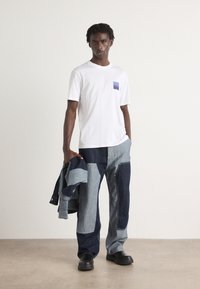 White t-shirt with a blue square pocket, paired with blue denim patchwork pants. A dark denim jacket is draped over one arm. Black shoes.