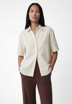 Kobieta o długich czarnych włosach, ubrana w luźną beżową koszulę na krótki rękaw zapinaną na guziki oraz brązowe prążkowane spodnie, ręce w kieszeniach, neutralne tło.