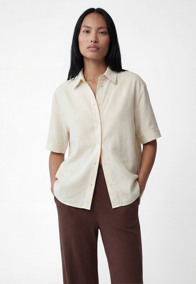 Mujer con cabello largo y negro, vestida con una camisa beige holgada de manga corta y pantalones marrones acanalados, con las manos en los bolsillos, fondo neutro.