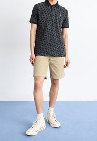 Personne portant un polo noir à motifs, un short beige, des chaussettes blanches, et des baskets montantes crème se tenant sur un sol bleu clair.