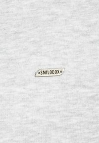Szara bawełniana koszulka z małą, srebrną metką z napisem "SMILODOX", wszytą po lewej dolnej stronie. Miękka w dotyku.