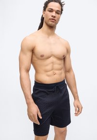 Homme torse nu avec des dreadlocks portant un short noir à cordon, debout devant un fond clair uni.