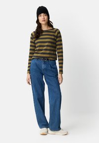 Langärmliges oliv- und senffarben gestreiftes Hemd, kombiniert mit locker sitzender blauer Jeans und weißen Turnschuhen, getragen mit einer schwarzen Mütze.