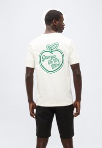 Άνδρας με την πλάτη προς τα εμπρός, φοράει λευκό μπλουζάκι με πράσινο περίγραμμα ροδάκινου και κείμενο "Georgia On My Mind" στην πλάτη, μαύρα σορτς, στέκεται μπροστά σε λευκό φόντο.