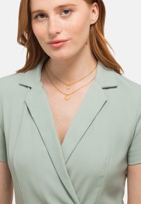 Abito avvolgente verde chiaro con maniche corte, caratterizzato da una scollatura a V e una texture sottile. Due collane circolari dorate sovrapposte al collo.