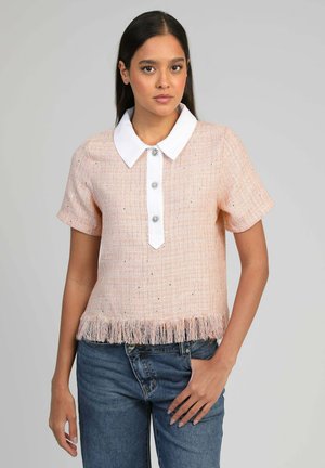 Femme portant un haut rose clair à manches courtes avec col blanc, trois boutons décoratifs, ourlet à franges, et un jean bleu sur un fond uni.