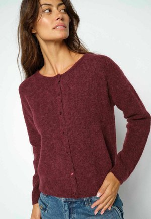 Woman wearing a buttoned, long-sleeve maroon cardigan and blue jeans, looking off to the side against a plain white background.