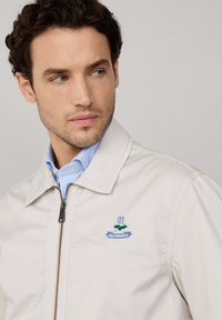 Hombre con cabello rizado corto que lleva una chaqueta beige con cremallera sobre una camisa de cuello azul claro, mirando hacia la izquierda.