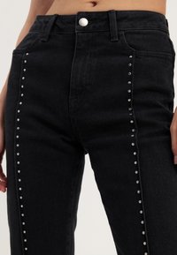 Sorte denimjeans med høy midje og sølv nagler langs sidesømmene. Stoffet har en glatt tekstur og litt stretch.