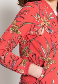 Chaqueta roja con un patrón floral en amarillo y verde, que cuenta con una cremallera y un bolsillo. La tela tiene una apariencia texturizada y puños de canalé.