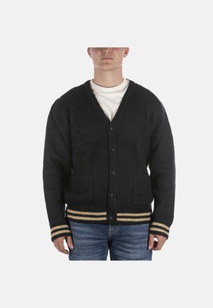 Man draagt een zwarte gebuttonde cardigan met beige streepaccenten op de manchetten en zoom over een wit overhemd en blauwe spijkerbroek, staand frontaal.