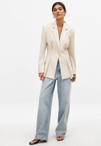 Beiger maßgeschneiderter Blazer mit Spitzklappen und einem Knopf, kombiniert mit hellblauen, weit geschnittenen Jeans und braunen Sandalen. Minimalistisches Design.