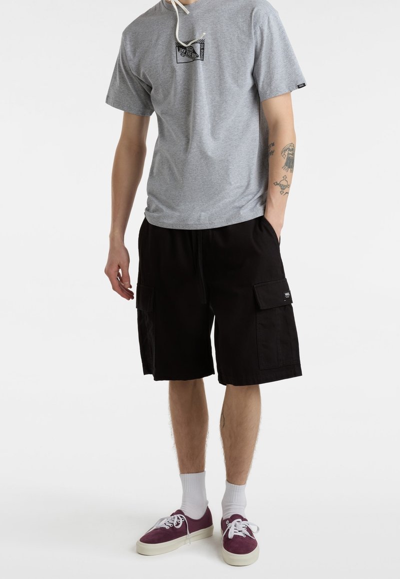 Homme portant un t-shirt Vans gris, un short cargo noir, des chaussettes blanches et des baskets bordeaux, debout avec une main dans la poche.