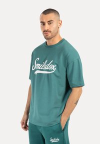 Camiseta de algodón en color verde azulado con cuello redondo y mangas cortas, que presenta un gran logotipo en letra cursiva blanca en el pecho.