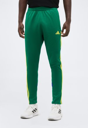 Mand iført grønne sportssbukser med gule sidelommer og hvide sneakers, stående mod en ensartet lys baggrund.