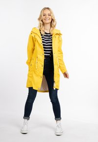 Cască galbenă impermeabilă cu glugă, nasturi frontali cu capse și buzunare laterale, asortată cu un tricou cu dungi și blugi negri.