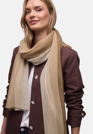 Nainen, joka pukeutunut pitkään, beigeen ja ruskeaan ribbineulottuun huiviin ruskean napillisen takin ja valkoisen paidan päällä, seisomassa yksiväristä taustaa vasten.