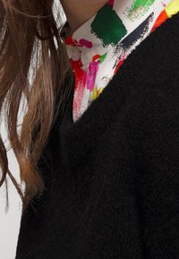 Gros plan d’une personne portant un pull noir texturé par-dessus une chemise blanche avec des motifs de taches de peinture colorées et de longs cheveux bruns.