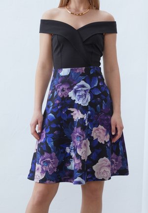 Kobieta ubrana w czarną sukienkę z odkrytymi ramionami, z kwiecistą spódnicą w odcieniach fioletu, niebieskiego i bieli, uzupełniona złotym naszyjnikiem.