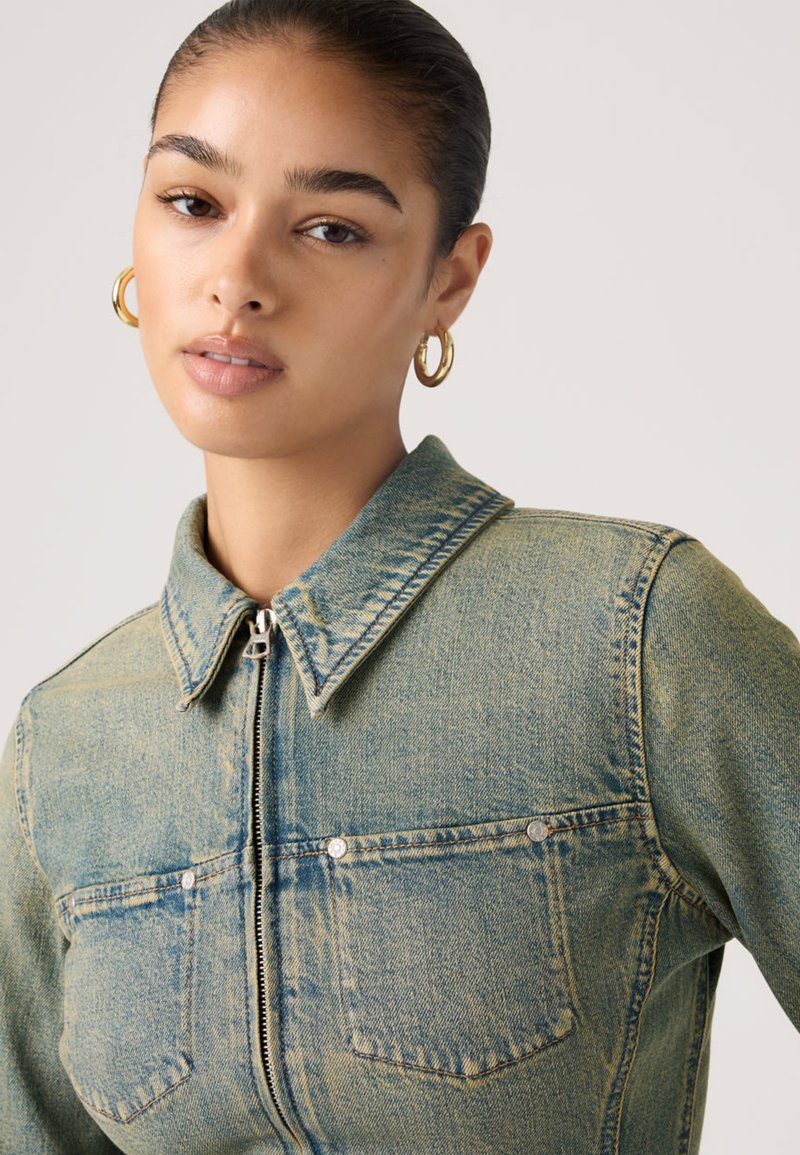 Femme aux cheveux tirés en arrière, portant des boucles d'oreilles en or en forme d'anneau et une veste en denim délavé avec fermeture éclair et poches poitrine, sur un fond uni.