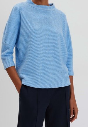 Persoon draagt een lichtblauwe geribde trui met driekwart mouwen en donkerblauwe broek, staand tegen een effen lichte achtergrond.