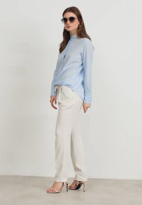 Camicia azzurro chiaro a maniche lunghe, pantaloni bianchi su misura e sandali con tacco trasparente; il modello indossa occhiali da sole scuri e ha i capelli mossi.