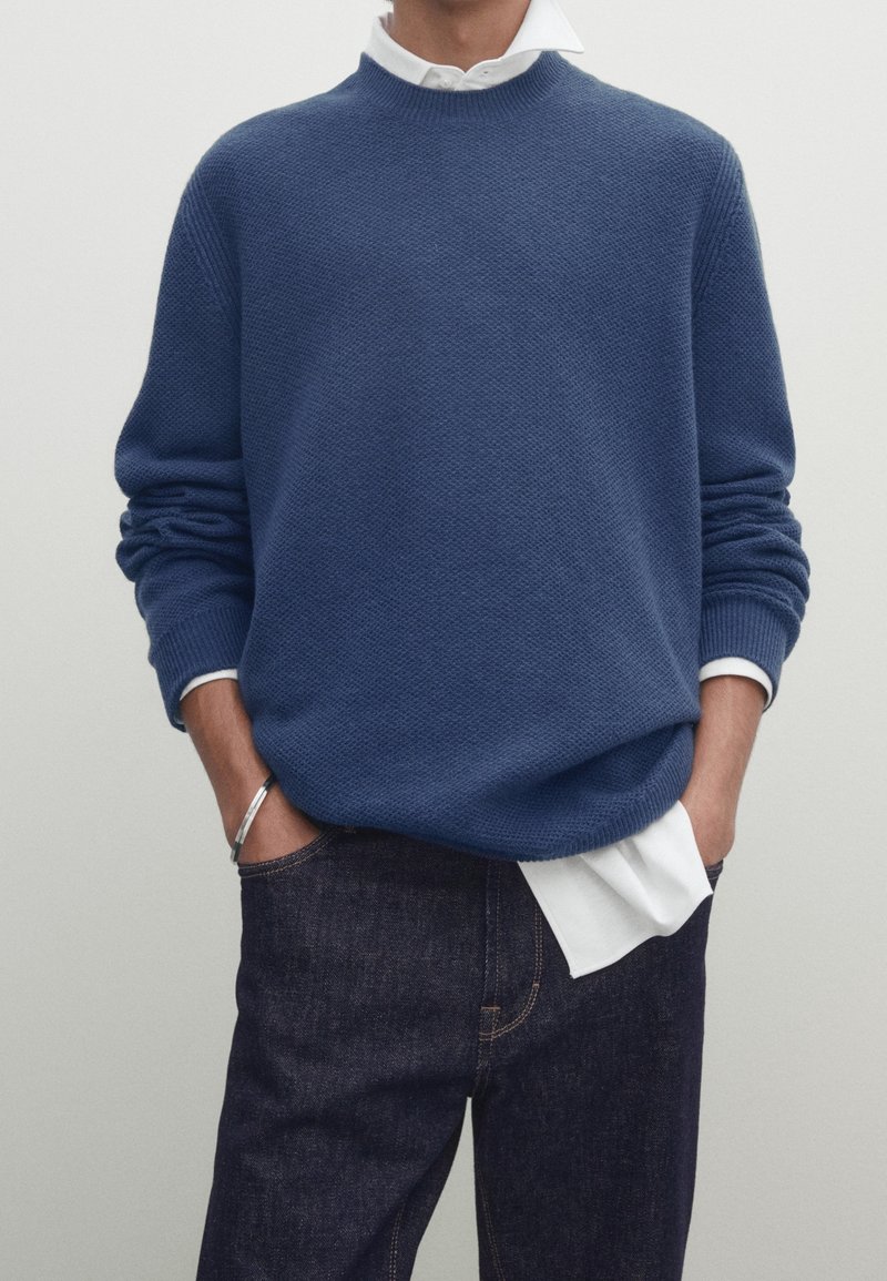 Pull en tricot bleu avec un motif texturé, col rond et épaules tombantes, porté sur une chemise blanche et associé à un jean foncé.