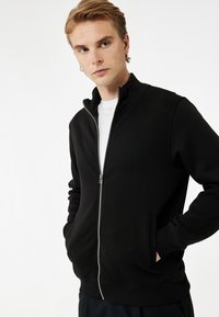 Chaqueta negra de cierre completo con alto cuello, bolsillos frontales, puños acanalados y una textura suave. Lleva puesta una camisa blanca.