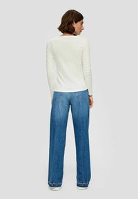 Weißes Langarmshirt und hellblaue weite Jeans mit aufgesetzten Taschen und lässiger Passform, vor einem grauen Hintergrund.