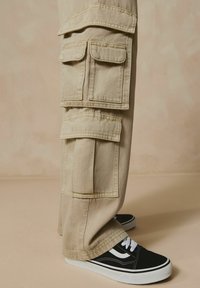 Pantalons cargo beiges avec plusieurs poches à rabat, tissu texturé et coupe décontractée, associés à des baskets noires avec des accents et des lacets blancs.