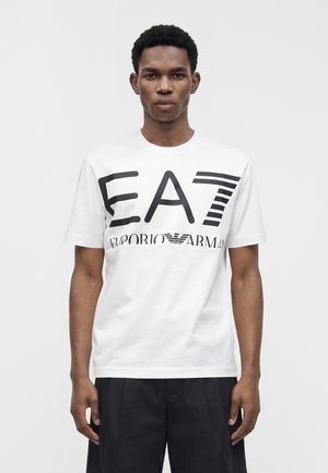 Jeune homme portant un t-shirt blanc avec un grand texte noir « EA7 Emporio Armani », debout devant un fond clair uni.