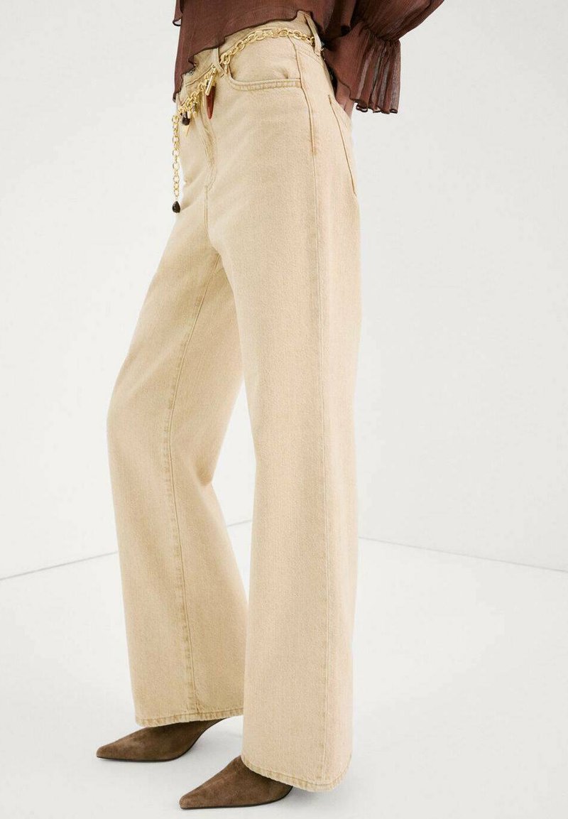 Pantaloni beige a gamba larga con tasche frontali, indossati con scarpe marroni a punta e una maglia marrone, accessoriati con una cintura a catena dorata.