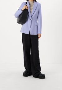 Donna che indossa un blazer lilla, un dolcevita beige, pantaloni neri larghi, scarpe con plateau e porta una borsa a spalla nera.