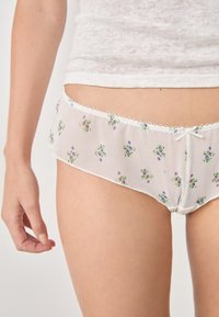 Las braguitas blancas de tela transparente presentan un patrón floral con pequeñas flores moradas y verdes. Un delicado borde de encaje y un lazo adornan la cintura.