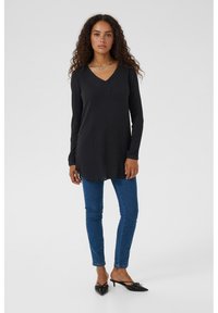 Black long-sleeve tunic with a V-neck and front placket, paired with blue skinny jeans and black heeled shoes. Simple texture, minimal design.