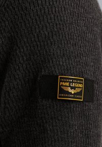 Dunkelgrauer, strukturierter Pullover mit einem Rippmuster. Verfügt über ein schwarzes Etikett mit goldener Schrift und grafischen Details, die Marke und Stil anzeigen.