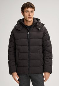 Giovane uomo vestito con un giubbotto imbottito nero con cappuccio, in piedi rivolto in avanti contro uno sfondo neutro.