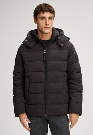 Junger Mann in einer schwarzen gepolsterten Jacke mit Kapuze, der mit dem Gesicht nach vorne vor einem schlichten Hintergrund steht.