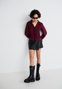 Βουργουνδίνη μπλούζα με ρυτίδες και μακριά μανίκια, συνδυασμένη με μαύρη μίνι φούστα από ψεύτικο δέρμα και χοντρές μαύρες μποτάκια με κορδόνια.