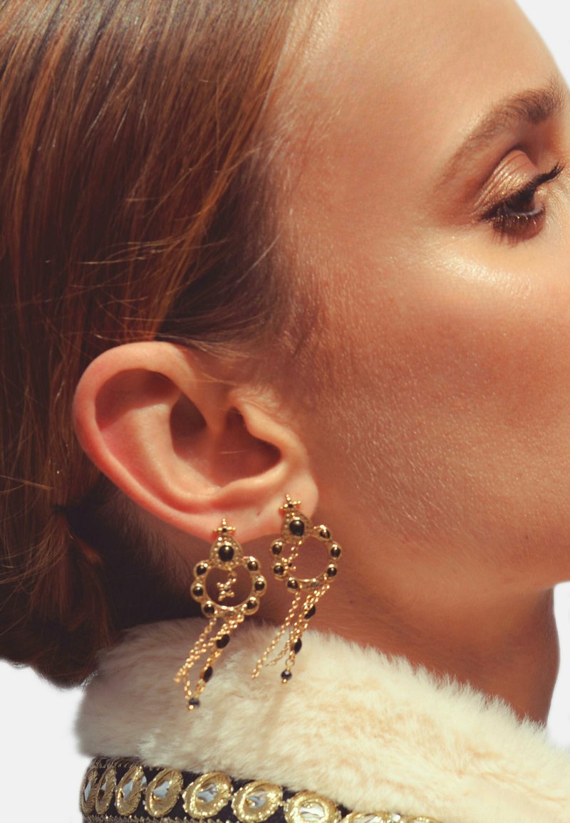 Boucles d'oreilles en or avec des motifs circulaires, ornées de perles noires et de chaînes en cascade. L'arrière-plan est flou, mettant l'accent sur les bijoux.