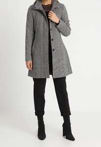 Donna che indossa un cappotto grigio a quadri di lunghezza al ginocchio sopra un top in pizzo nero, pantaloni neri con risvolto e stivaletti neri con tacco, in piedi su uno sfondo bianco.