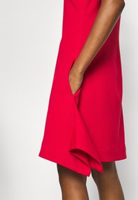 Röd klänning av glatt tyg, med en sidficka och en rak fåll. Designen är utan ärmar med en figurnära siluett.