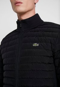 En närbild av en man som bär en svart puffjacka med hög krage, som har en liten grön och vit krokodil-logotyp på bröstet.