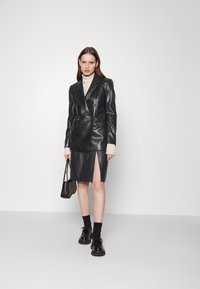 Zwarte faux leren blazer met een dubbele knoopsluiting, gecombineerd met een bijpassende rok met een zijsplit. Het model houdt een kleine handtas vast.