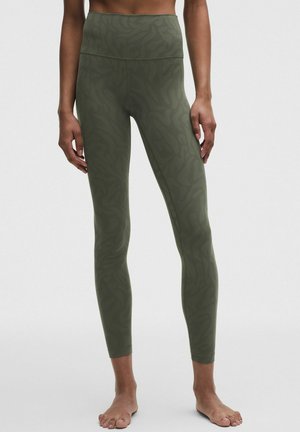 Vrouw draagt olijfgroene leggings met hoge taille en een subtiel abstract patroon, staat blootsvoets tegen een effen achtergrond.