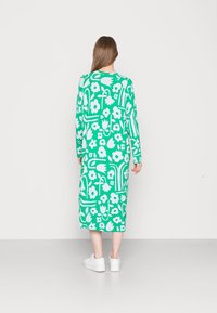 Vestido verde de mangas longas com padrões florais e abstratos brancos, comprimento até a metade da panturrilha, corte solto, combinado com ténis brancos.