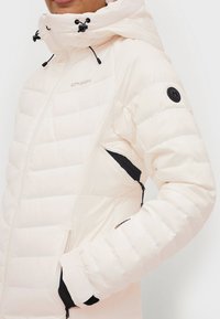 Kvinde iført en kremfarvet quiltet jakke med hætte, sidelomme med lynlås, logo-patch på ærmet, og hånd i frontlommen.