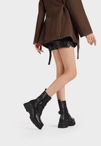 Stivali da combattimento in pelle nera con suola spessa, chiusura con lacci e dettaglio fibbia, e una texture liscia. Adatti per uno stile versatile.