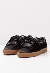 Pár černých semišových tenisek s hrubými tanovými gumovými podešvemi a širokými saténovými černými tkaničkami, položených vedle sebe na bílé ploše.