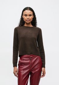 Donna con capelli ricci che indossa un maglione marrone scuro e pantaloni in pelle rossa a vita alta con cerniera frontale, in piedi davanti a uno sfondo bianco semplice.